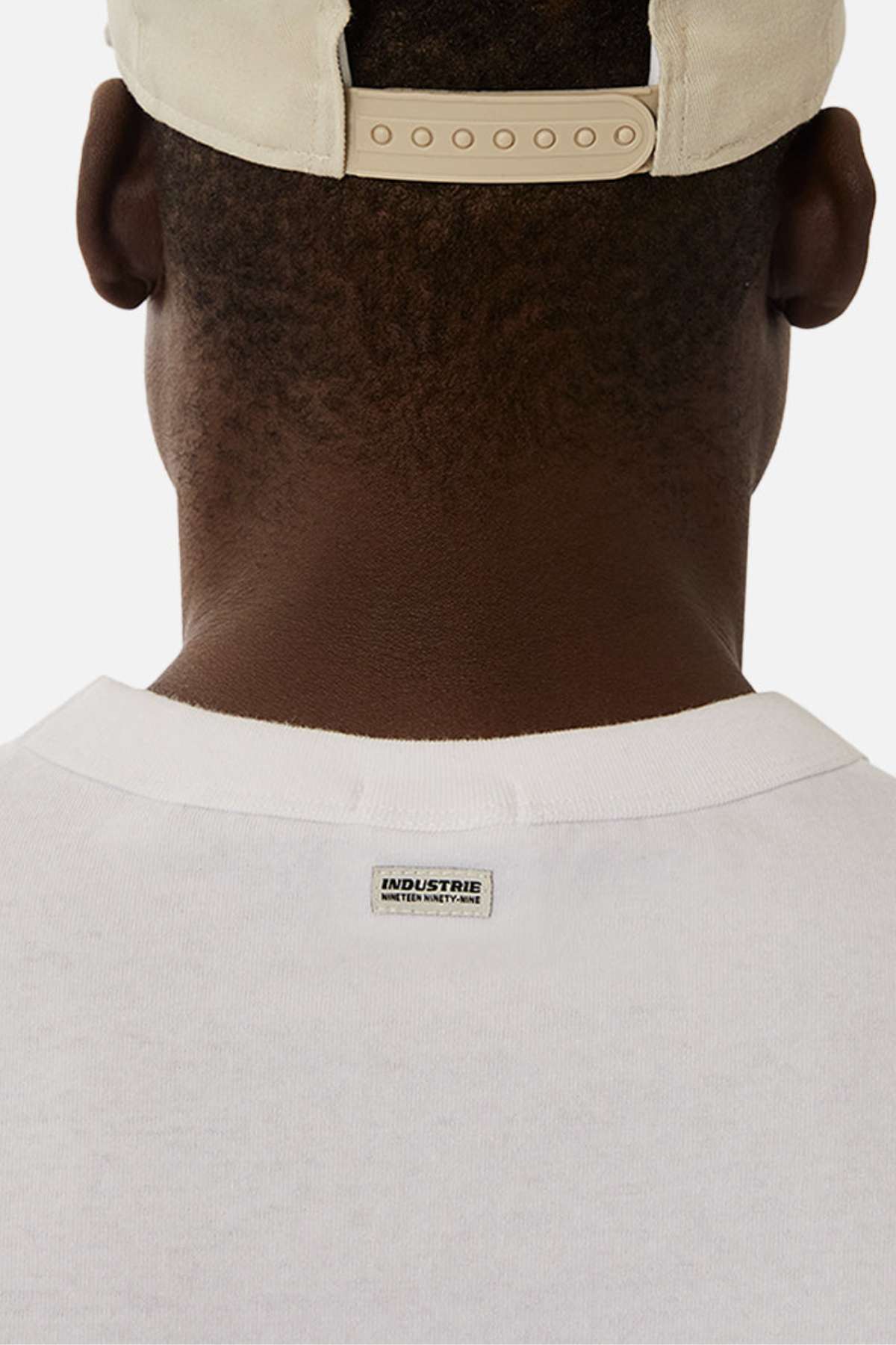 A close-up of the back of a person wearing the Industrie The Del Sur Sleeveless Tee in white, made from 100% cotton with an "INDUSTRIE" label on the collar and a beige baseball cap, set against a plain white background.