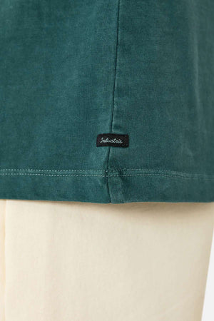 Close-up of the bottom hem of the Industrie The Del Sur Tee in green, featuring a small black "Industrie" tag. Worn over cream-colored pants and crafted from 100% cotton for comfort and style.