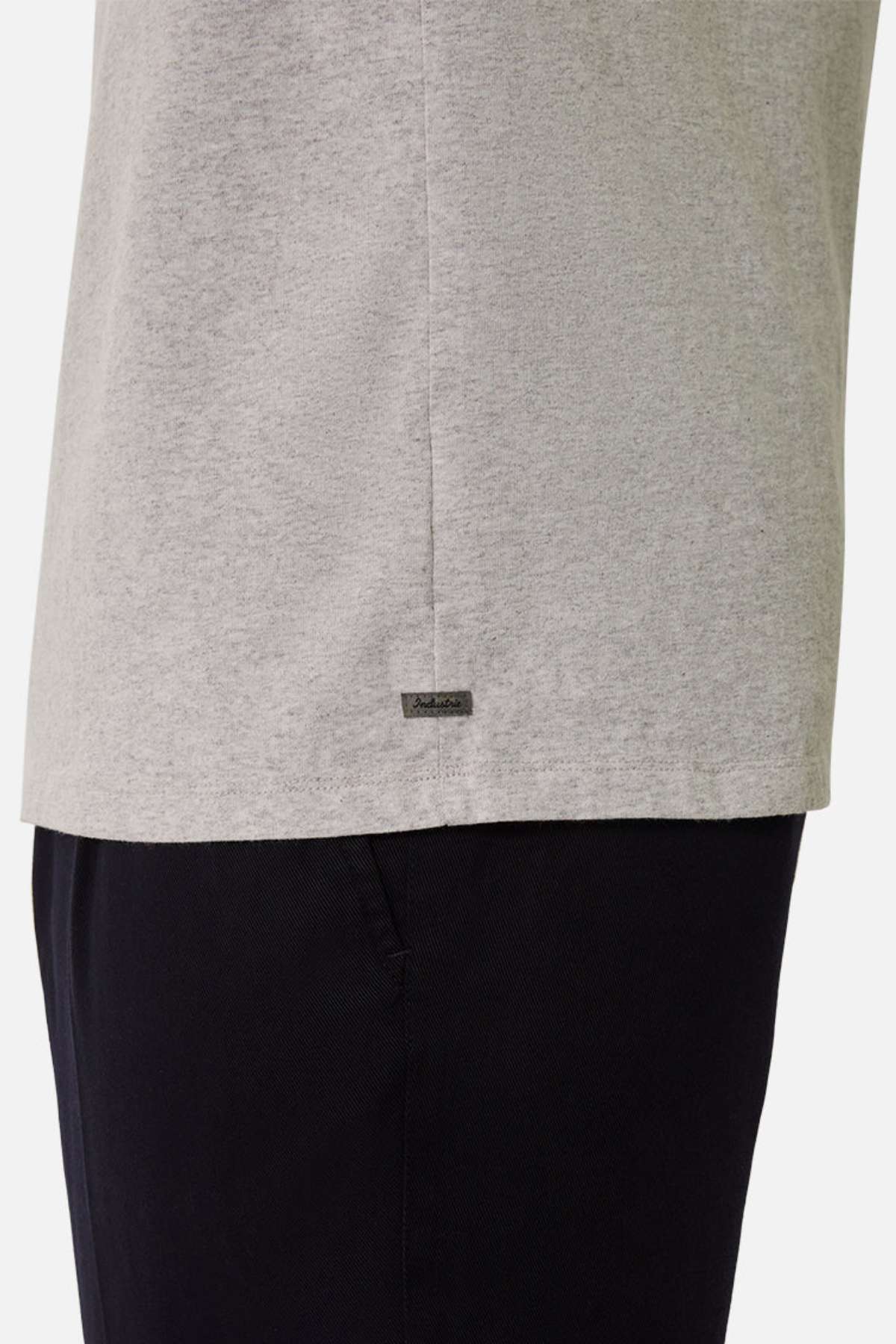 A close-up side view of a person wearing the Industrie The Del Sur Tee in light gray (100% cotton) and black pants. A small metal tag accents the hem. Set against a plain white background.