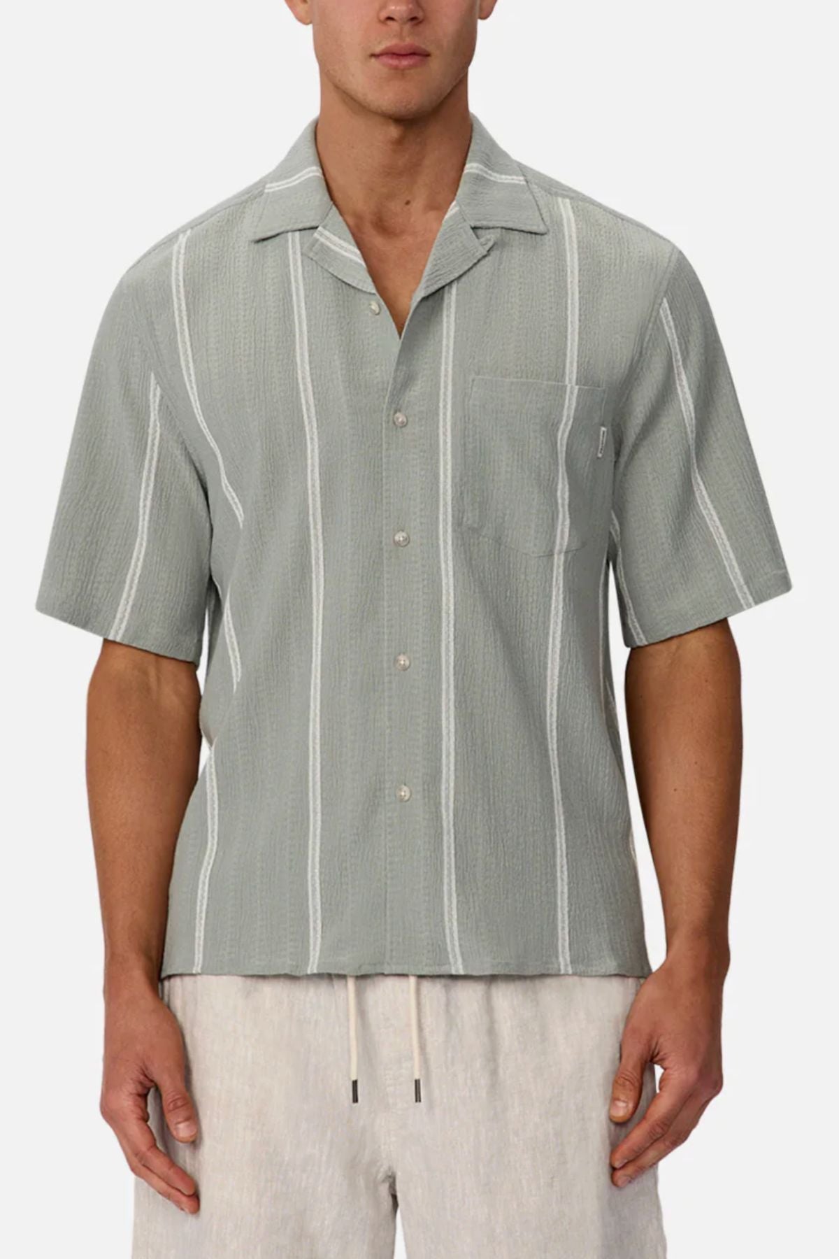 A man wears the Industrie The Fullerton Short Sleeve Shirt—light gray with vertical white stripes—paired with light beige drawstring shorts, standing against a plain white background.