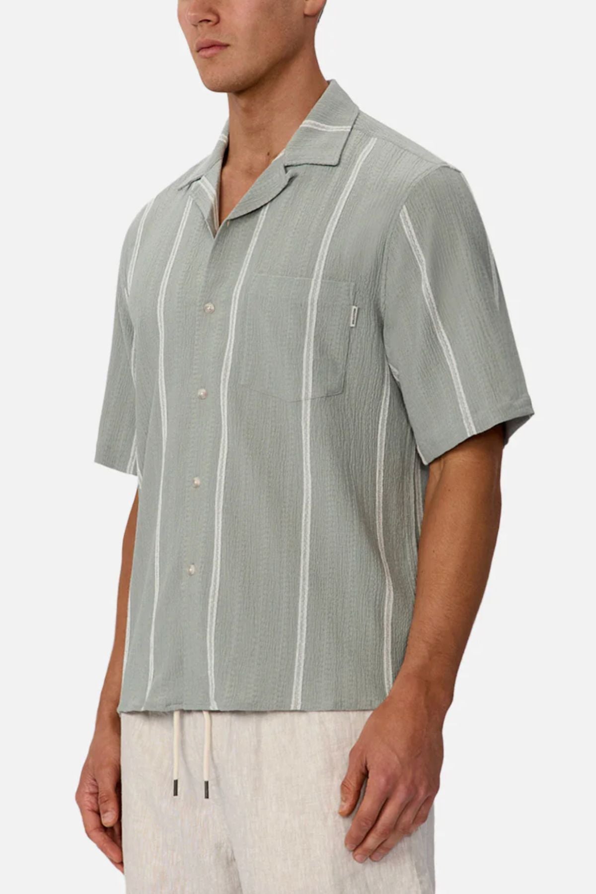 A man wears the Industrie The Fullerton Short Sleeve Shirt—a light gray, short-sleeve button-up with white stripes and a chest pocket—paired with light drawstring pants for a versatile look.