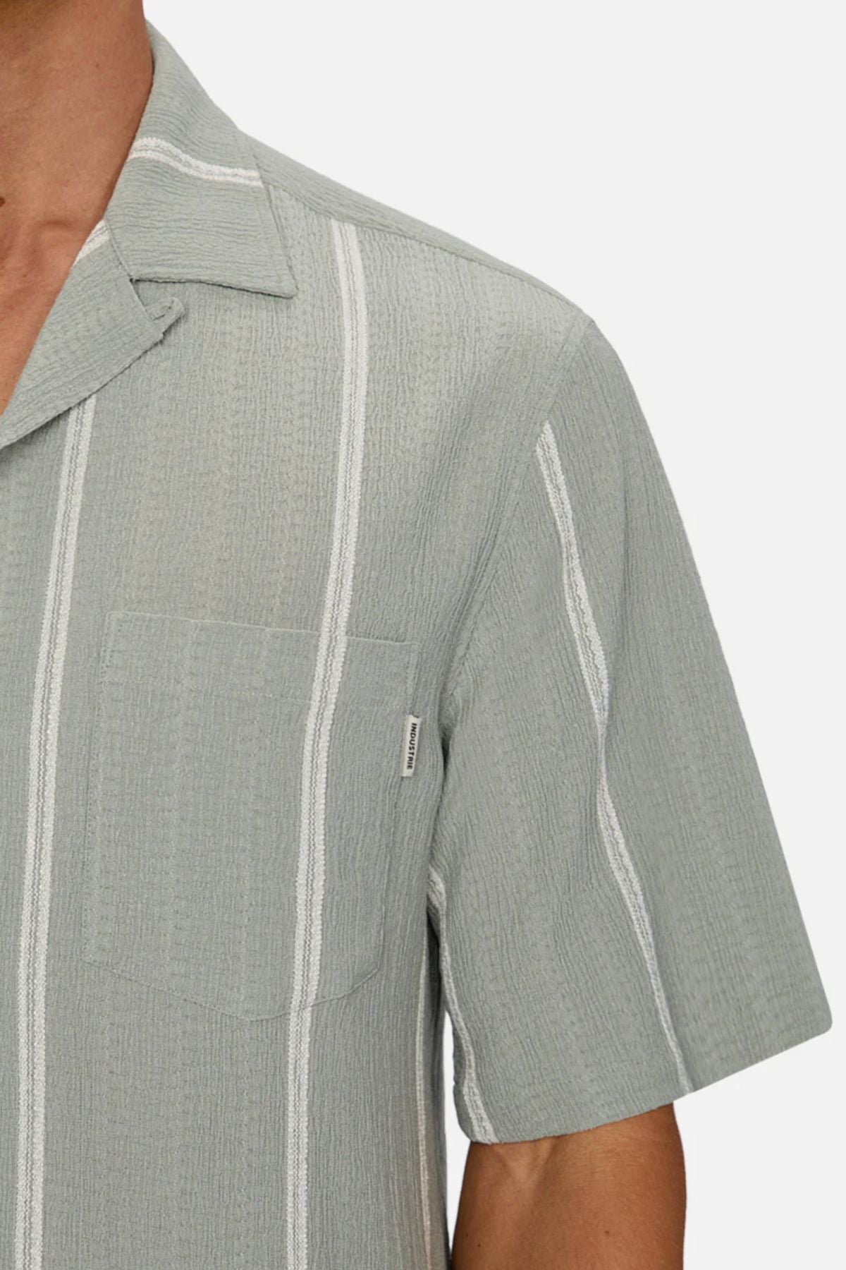 Close-up of a person wearing the Industrie The Fullerton Short Sleeve Shirt in light gray with vertical white stripes, short sleeves, chest pocket, and notched collar. Only the upper torso and part of the left arm are shown.