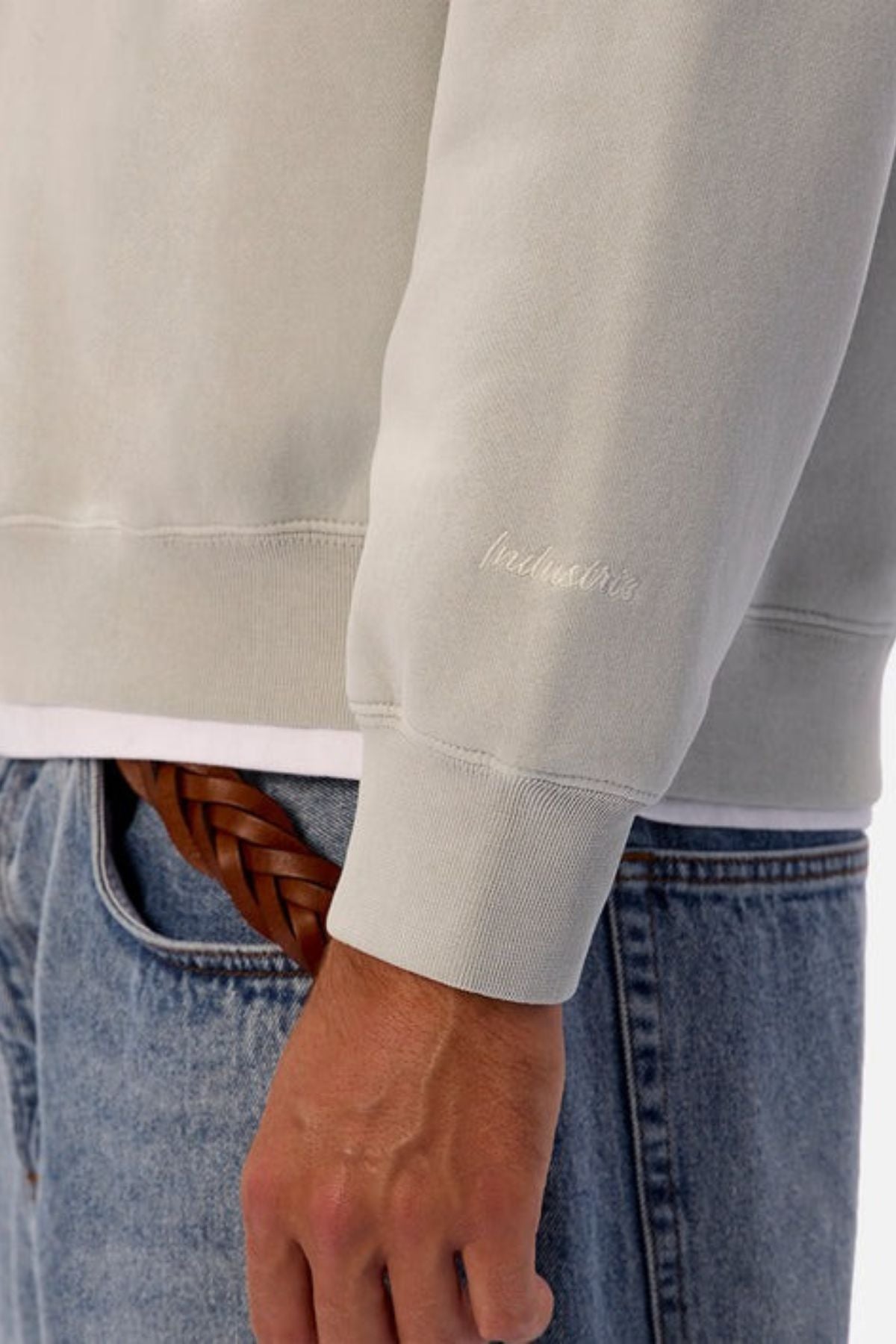 Someone is wearing the Industrie The New Del Sur Washed Sweat in an oversized fit with subtle wrist embroidery, paired with blue jeans, a brown braided belt, and a white shirt peeking out. Only their torso and arm are visible.
