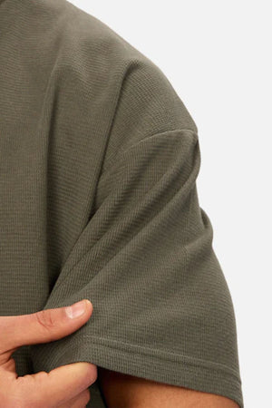 A close-up of a person wearing the Industrie The Marlon Tee in olive green waffle knit, gently pulling on the sleeve. The background is plain and light-colored.