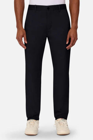 A person wears Industrie The Regular Cuba Chino Pant in black, paired with a white shirt and white sneakers, standing against a plain light background. The photo captures the look from shoulders to feet with arms relaxed at their sides.