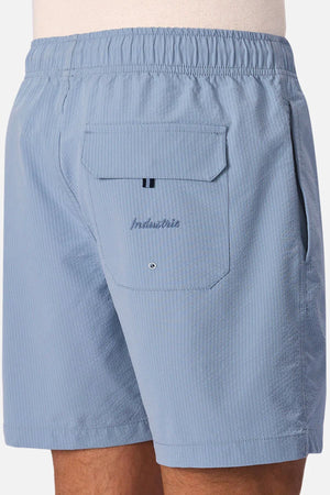 A close-up of a person wearing Industrie The New Seersucker Swim Short in light blue seersucker, featuring an elastic waistband and a back pocket with flap, grommet hole, and "Industrie" embroidery—crafted in quick-dry polyester.