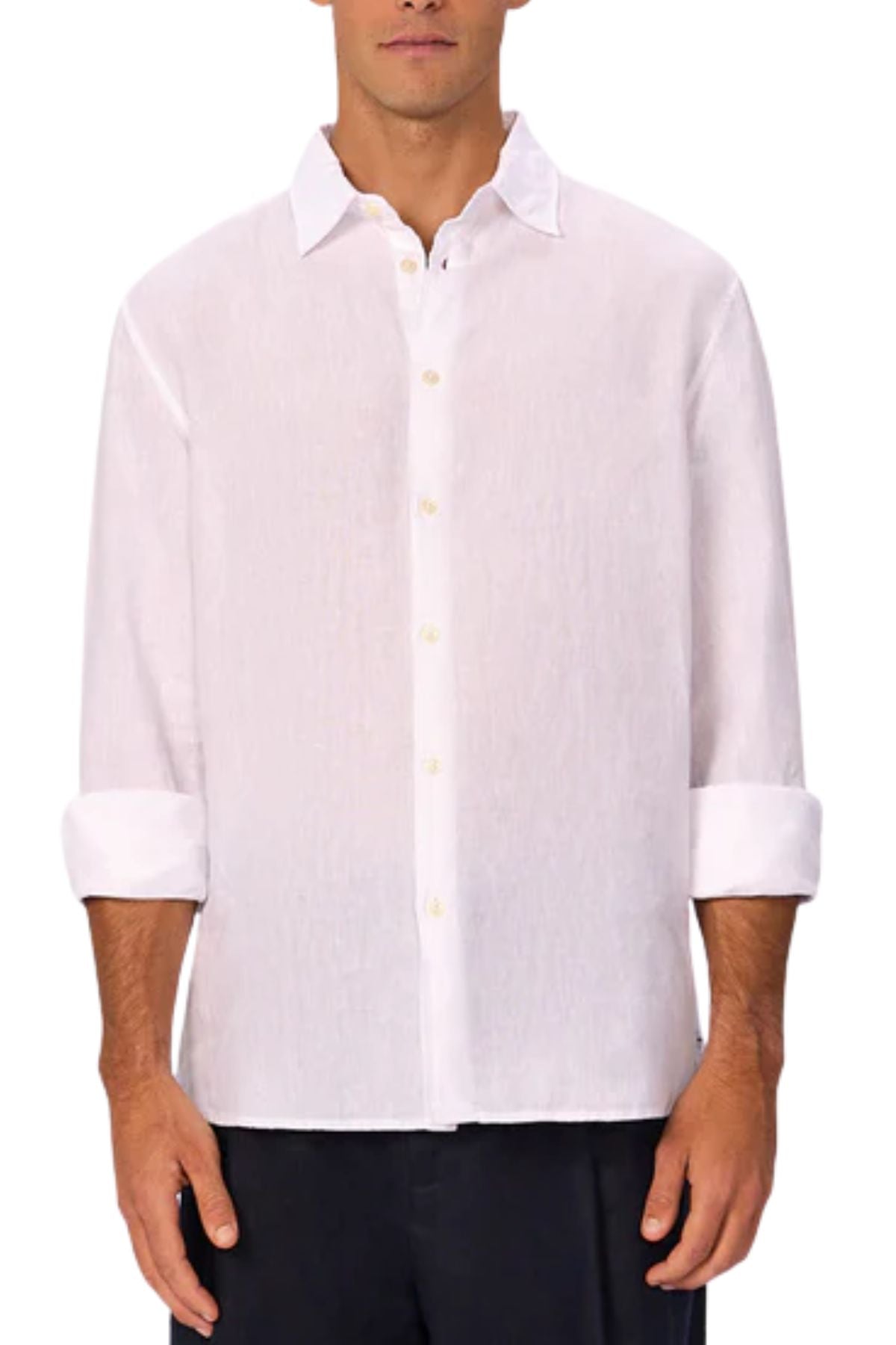 A man wears the Industrie The St Martins Shirt, a white long-sleeve linen shirt with rolled-up sleeves, untucked and paired with dark pants, standing against a plain background.
