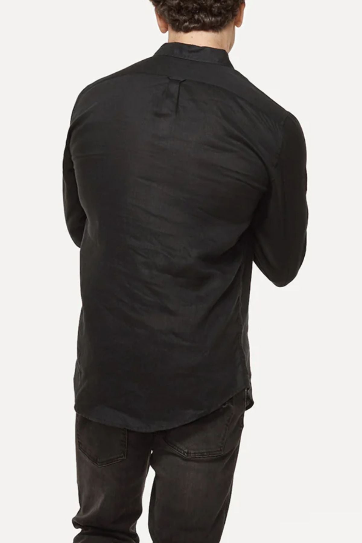 A man with curly dark hair faces away, wearing an Industrie The Tennyson Linen Shirt in a relaxed fit paired with dark jeans against a plain light background, his arms crossed.