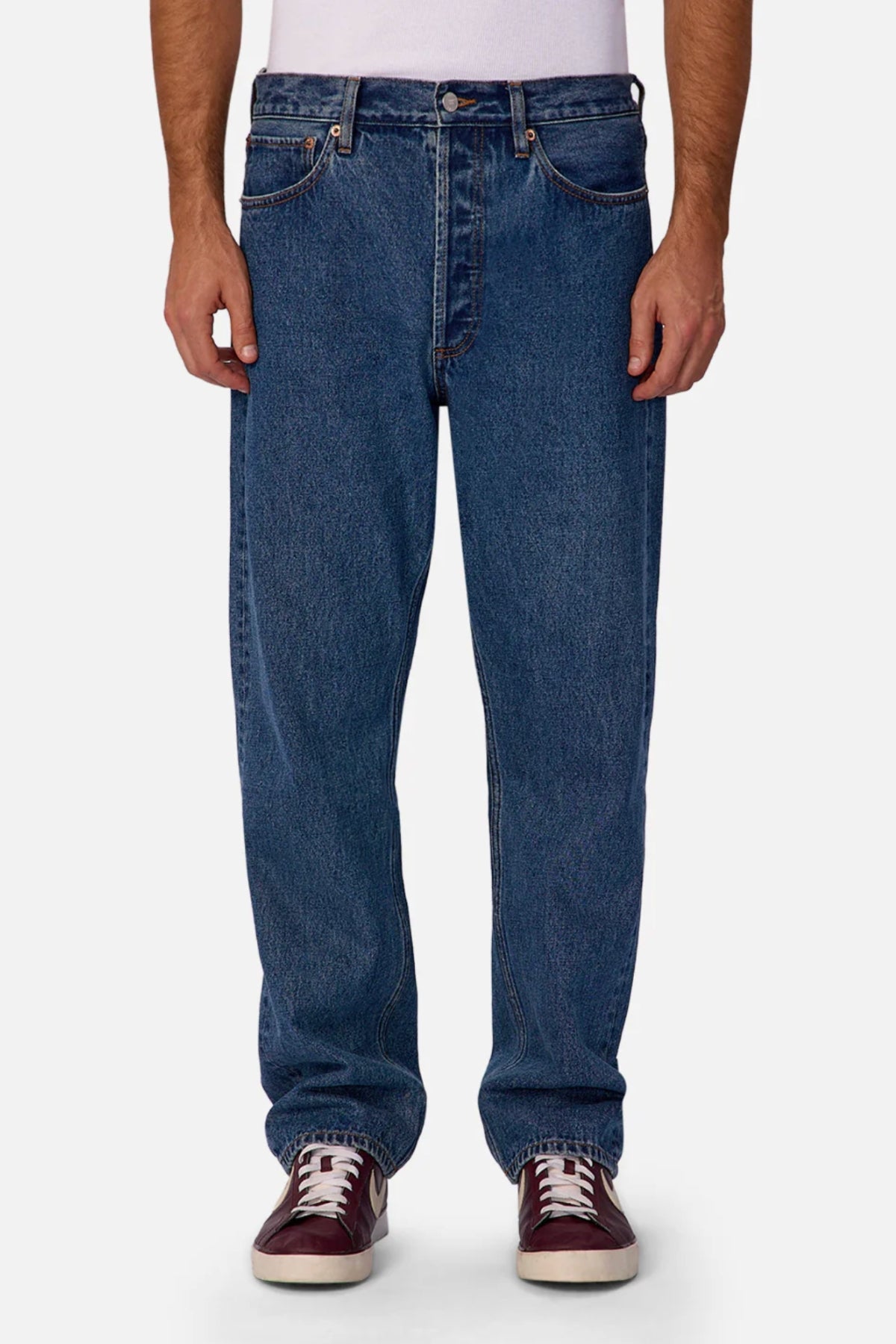 A person wearing Industrie The 90's Jean in vintage washed denim, a tucked-in white T-shirt, and maroon sneakers with white laces stands against a plain white background.