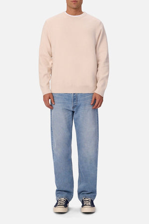 A man stands facing forward against a plain white background, wearing a light beige sweater, Industrie The 90's Jean in vintage washed denim, and black sneakers with white soles.