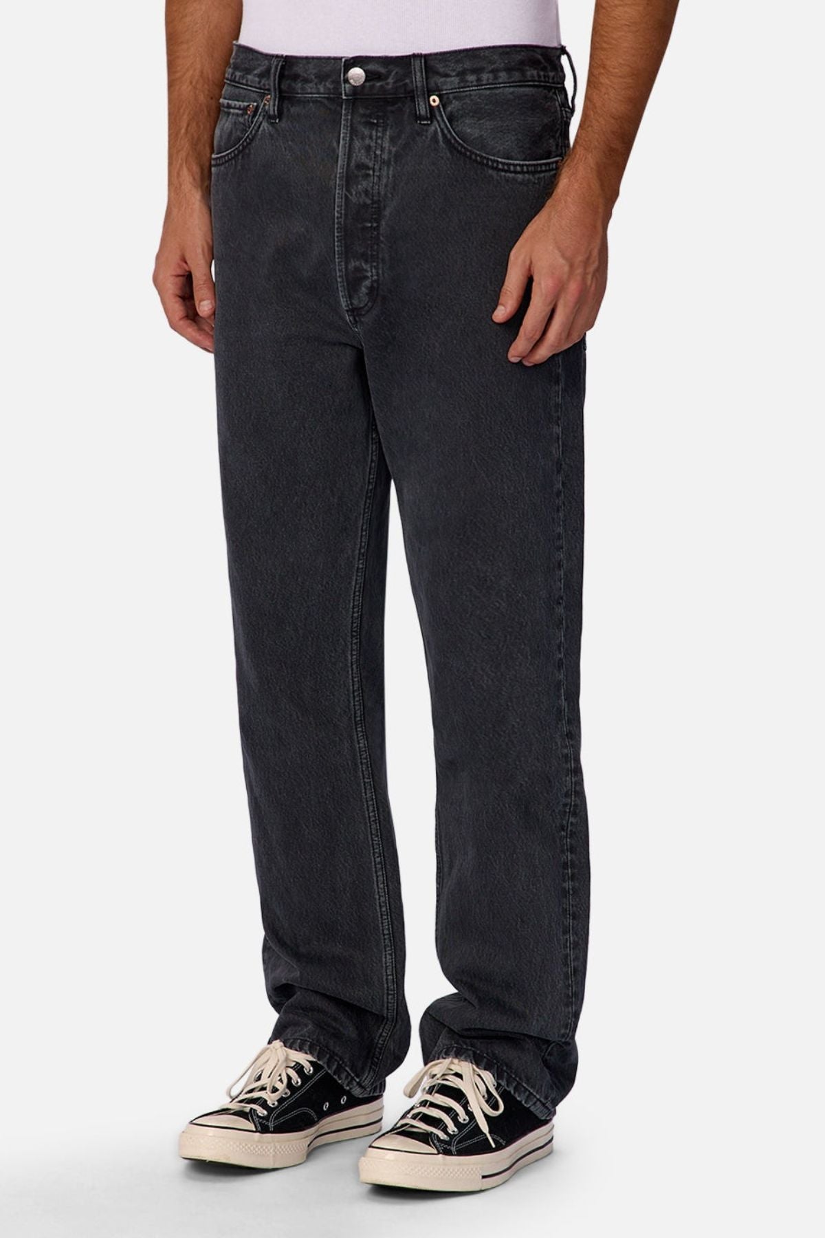 A person models Industrie The 90's Jean in vintage washed denim, paired with a white shirt and black-and-white lace-up sneakers against a plain white background, showing only their lower torso and legs.