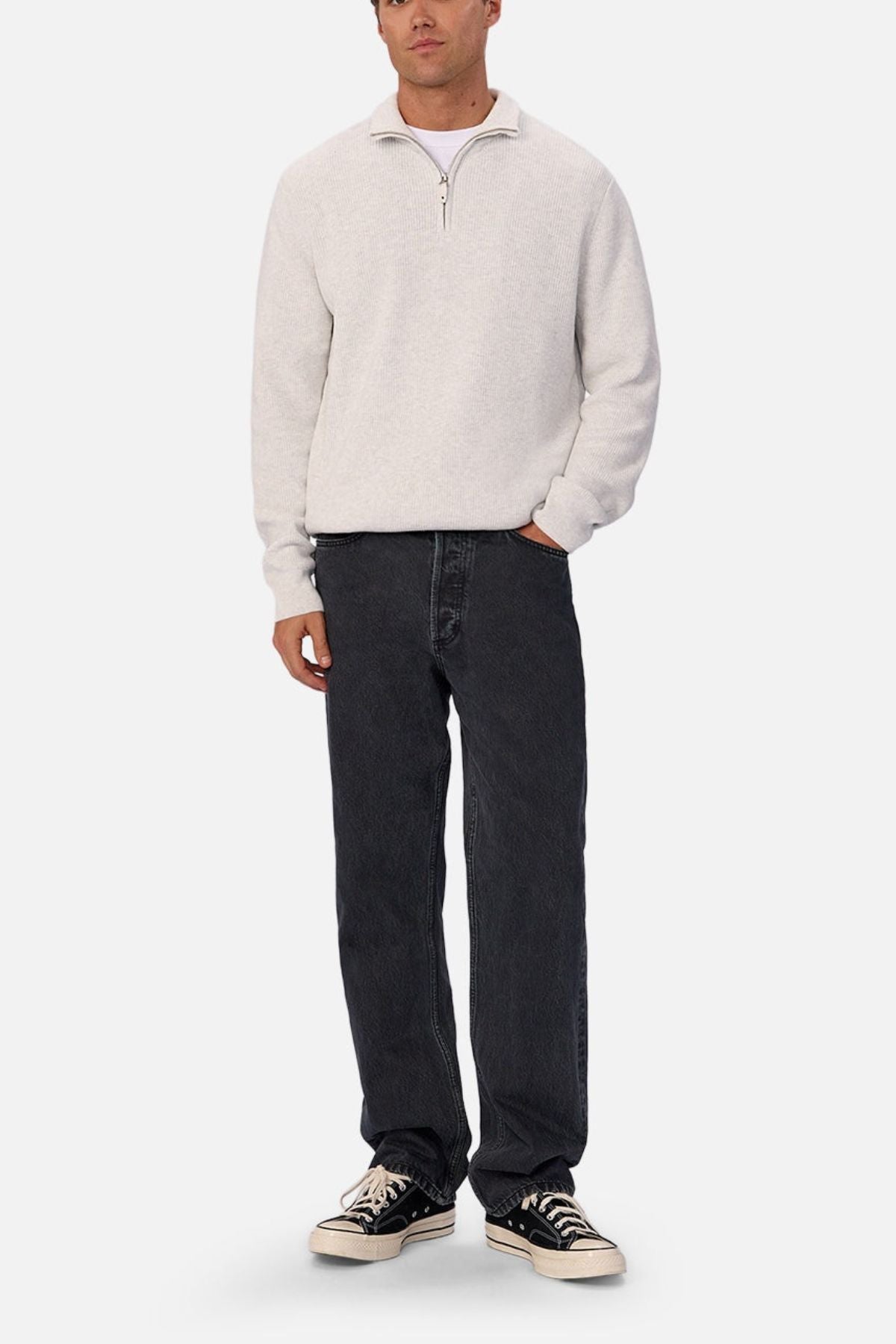 A person stands against a plain background wearing a light gray quarter-zip sweater, Industrie The 90's Jean in vintage-washed denim, and black low-top sneakers with white soles.
