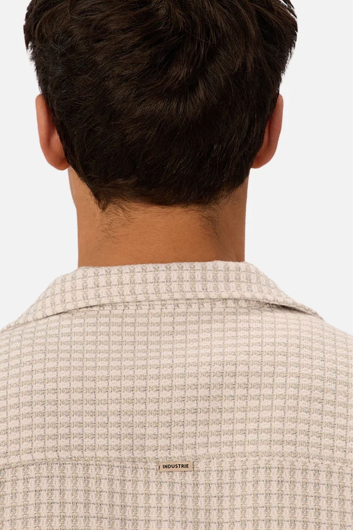 A person with short dark hair is seen from behind wearing the Industrie The Almeria Short Sleeve Shirt—a light-colored, textured cotton shirt featuring a small “INDUSTRIE” label on the upper back against a plain white background.