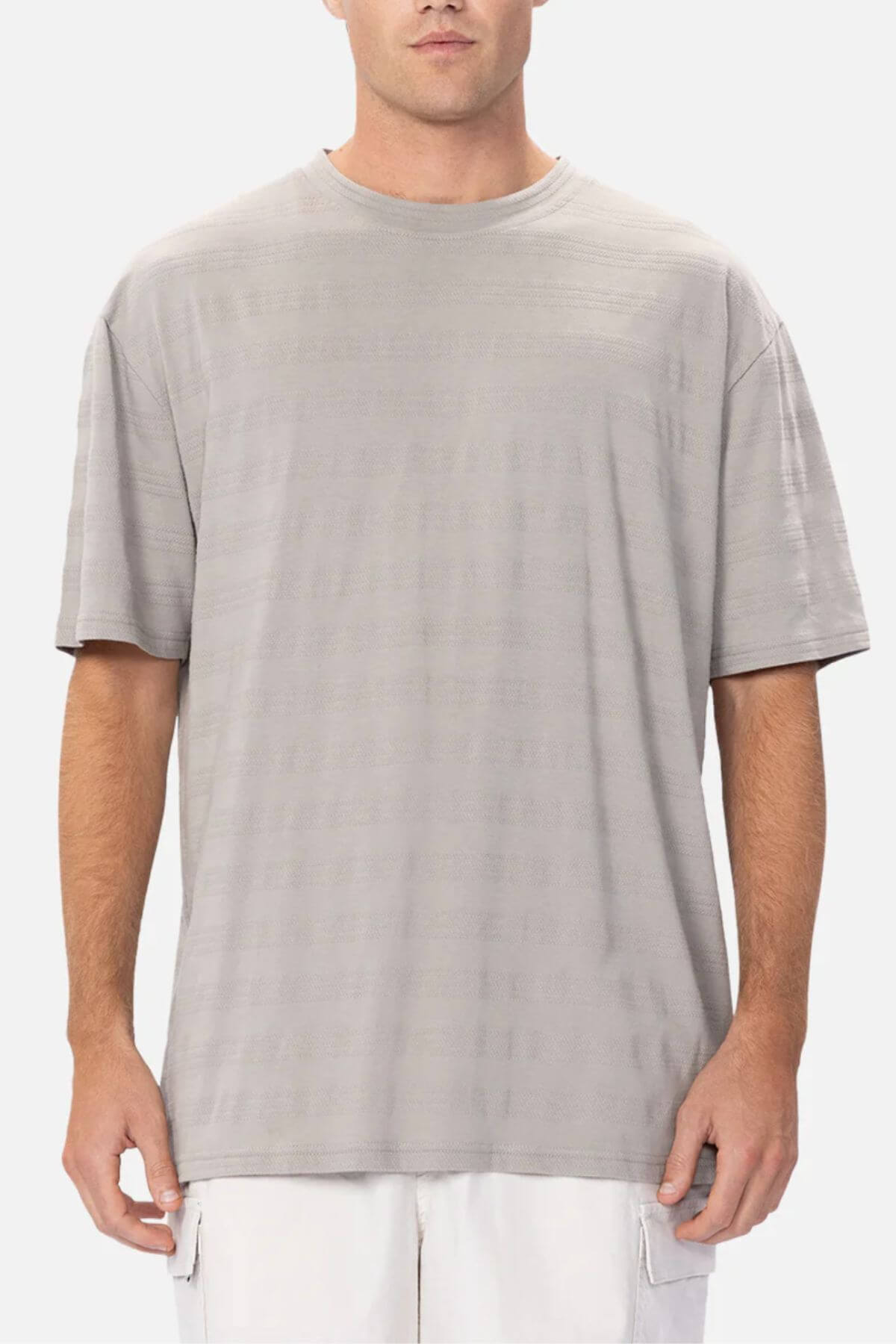 A man wears the Industrie The Anderson Textured Stripe Tee in light gray, featuring a loose fit, paired with white shorts as he stands facing forward against a plain light background.