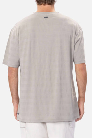 A person is seen from the back wearing the Industrie The Anderson Textured Stripe Tee in light grey with subtle horizontal stripes and a small logo tag near the neckline, paired with light shorts against a plain white background.