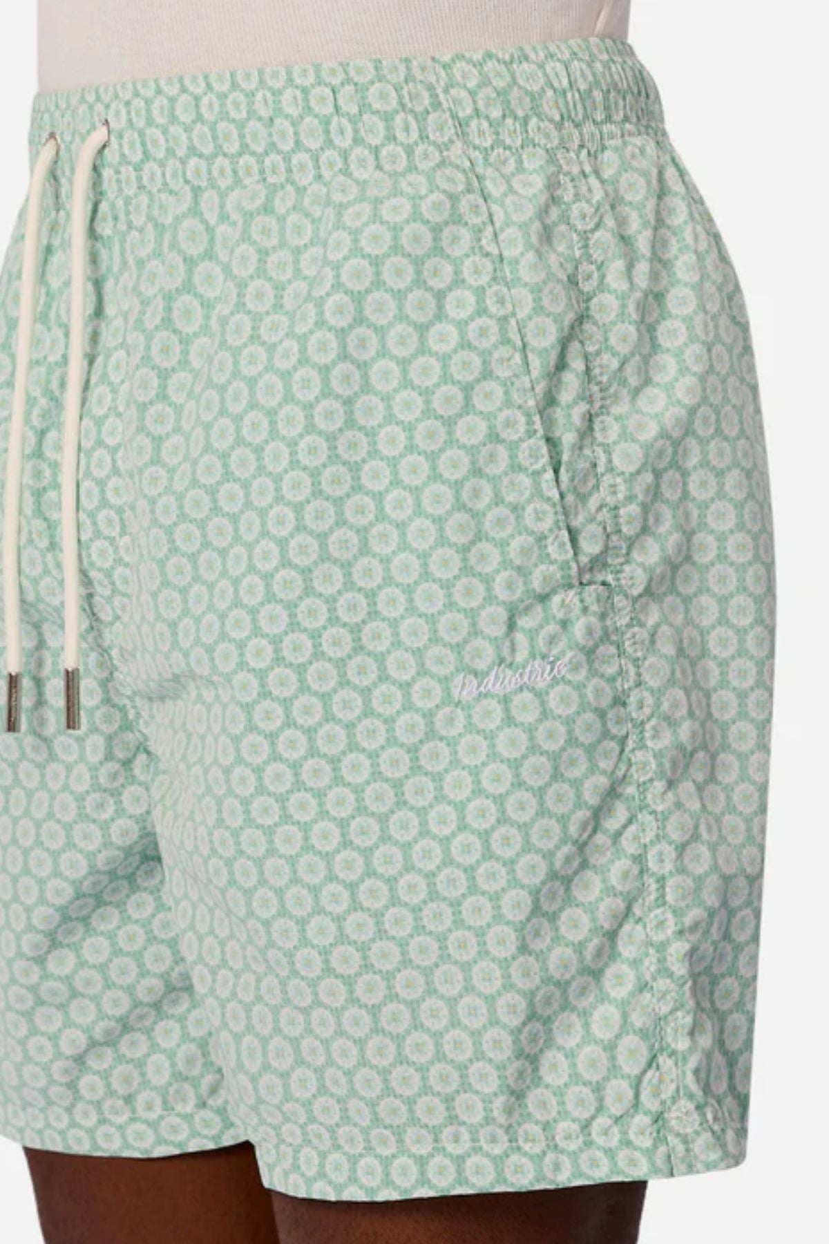 Someone wears the Industrie The Aruba Swim Short—light green men’s beach shorts with a white circular pattern, white drawstrings, a small embroidered logo on the left, quick-dry fabric, side pockets, and an elastic waistband.
