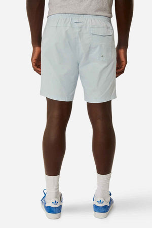 A person stands facing away, wearing light blue Industrie The Bahama Angler Shorts with a drawcord waist and a back pocket with a button, paired with a gray shirt, white socks, and blue Adidas sneakers. The background is plain white.