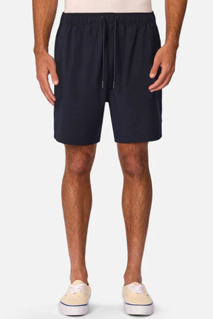 A person wears Industrie The Barbados Swim Short in navy blue, featuring an elastic waistband and drawstring. Beige slip-on shoes complete the look. Only the lower body is visible, with arms relaxed at their sides.