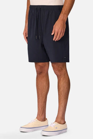A person wears Industrie The Barbados Swim Short in navy blue with light beige sneakers, standing against a plain white background—ideal men's beachwear for sunny getaways.