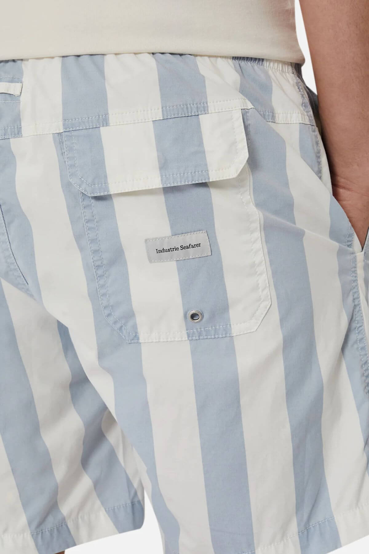 Close-up of a person wearing Industrie The Beverly Bahama Short in light blue and white vertical stripes, with a back flap pocket featuring an "Industrie Seafare" label. Made from breathable fabric for comfort, hand partially tucked into the pocket.