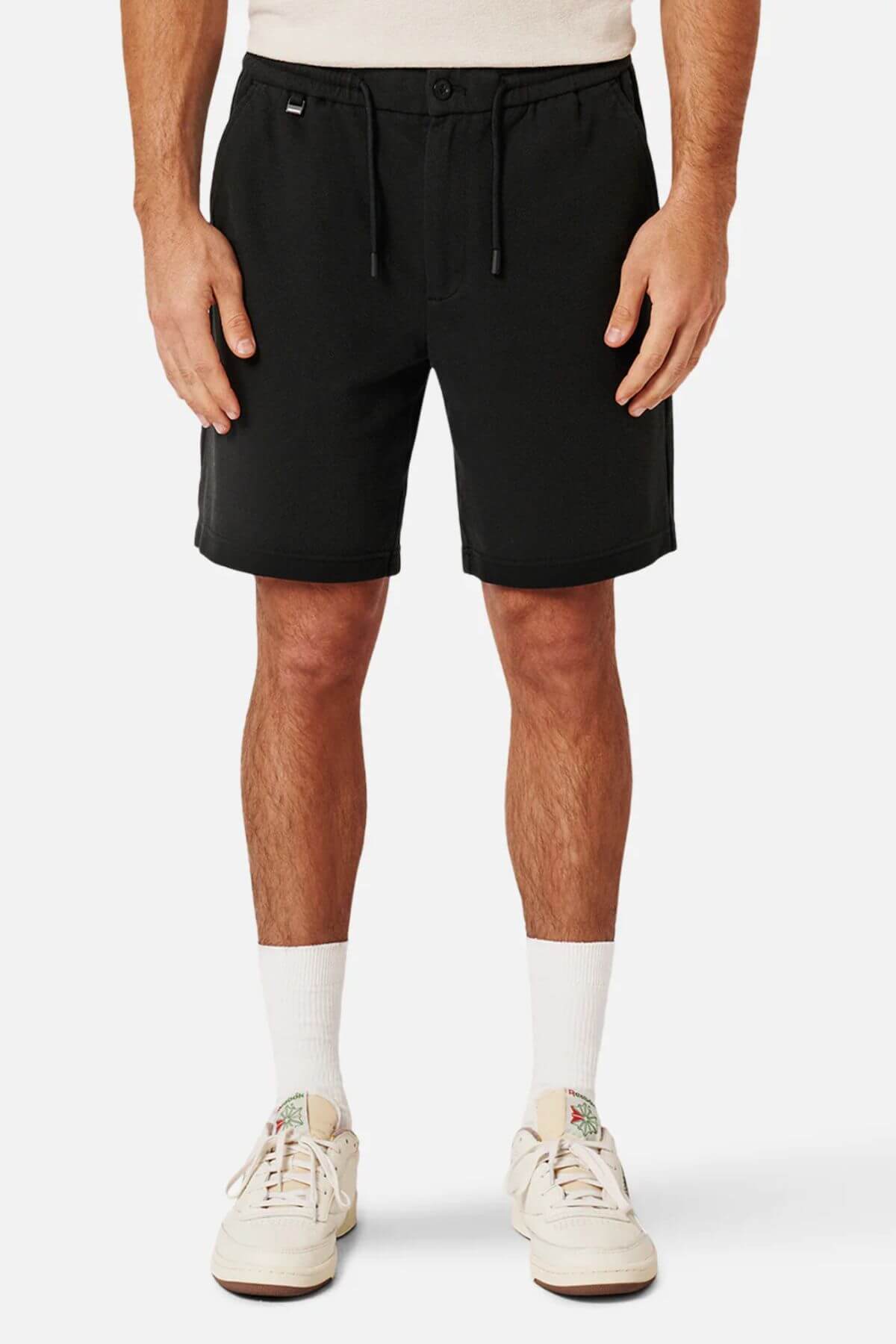 A person in Industrie The Broadway Short, styled in relaxed fit black, paired with white socks and sneakers, stands against a plain white background. Their hands rest comfortably by their sides.
