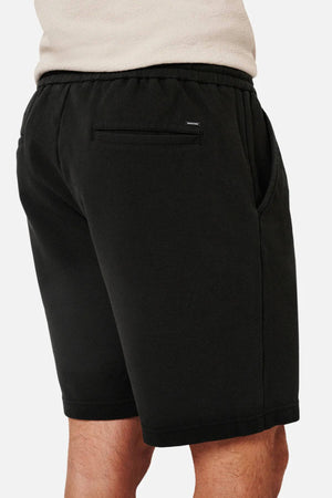 A person is seen from the side with their hand in the pocket of Industrie The Broadway Short, featuring black stretch fabric, a relaxed fit, an elastic waistband, and a small label on the back pocket. The scene is set against a plain white background.