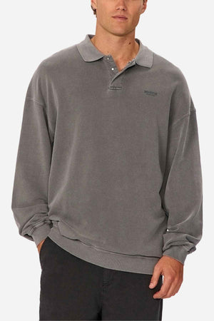 A person models the Industrie The Burbank Polo Track, a loose-fitting, long-sleeve gray collared shirt with buttons, paired with dark pants. Styled simply against a white background, one hand rests in their pocket.