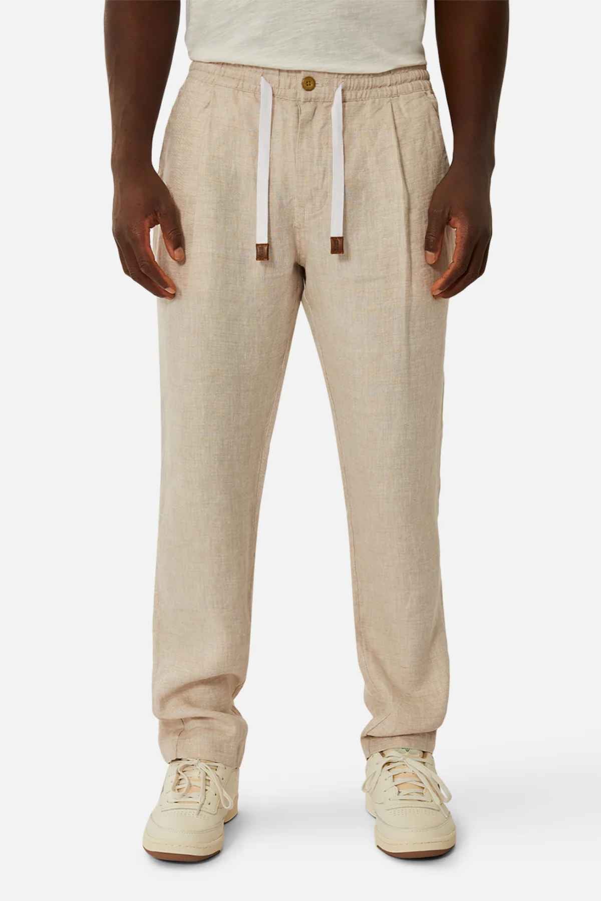 A person models the Industrie The Capri Linen Pant, styled in beige with a relaxed wide-leg fit and button closure, paired with white sneakers against a plain background. These summer-ready pants also feature a white drawstring.