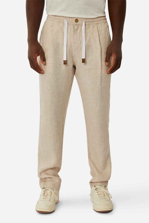 A person models the Industrie The Capri Linen Pant, styled in beige with a relaxed wide-leg fit and button closure, paired with white sneakers against a plain background. These summer-ready pants also feature a white drawstring.
