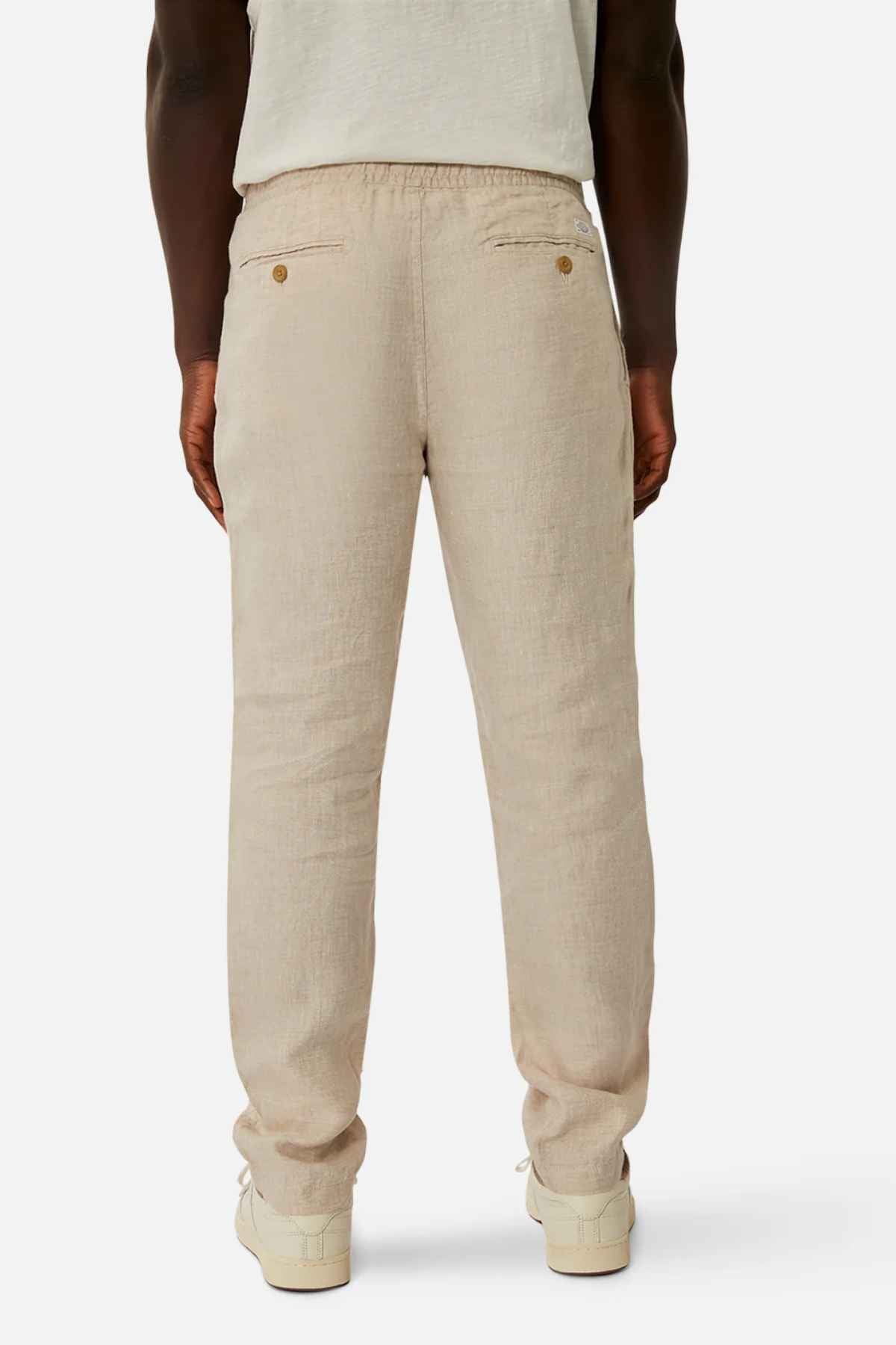 A person is seen from the back wearing Industrie The Capri Linen Pant in light beige, wide-leg fit with two buttoned back pockets, paired with a white shirt and off-white shoes against a plain white background, exuding a summer vibe.
