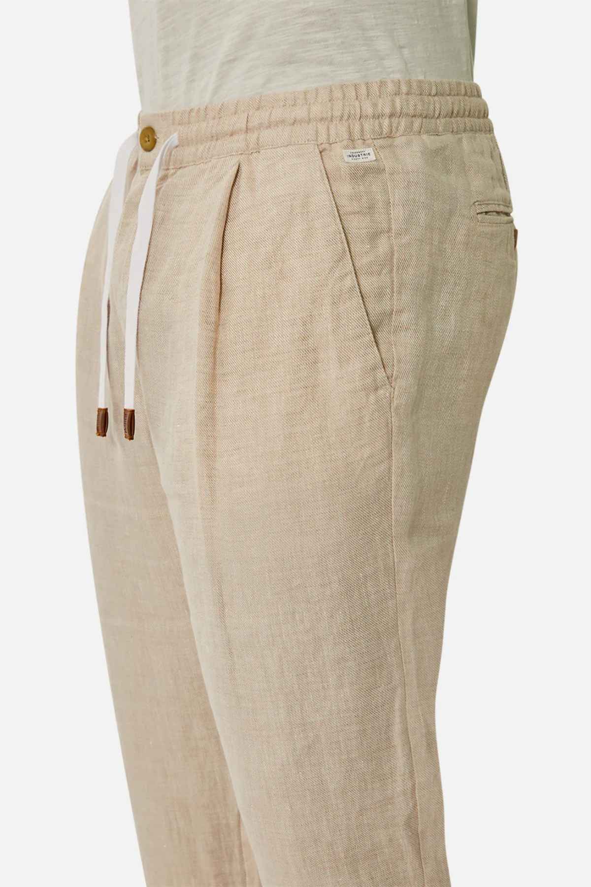 Side view of Industrie The Capri Linen Pant in beige, showcasing an elastic waistband with a white drawstring for a summer vibe. The pants feature pleats, a wooden button detail, and a wide-leg fit, paired effortlessly with a light cream shirt for a stylish look.