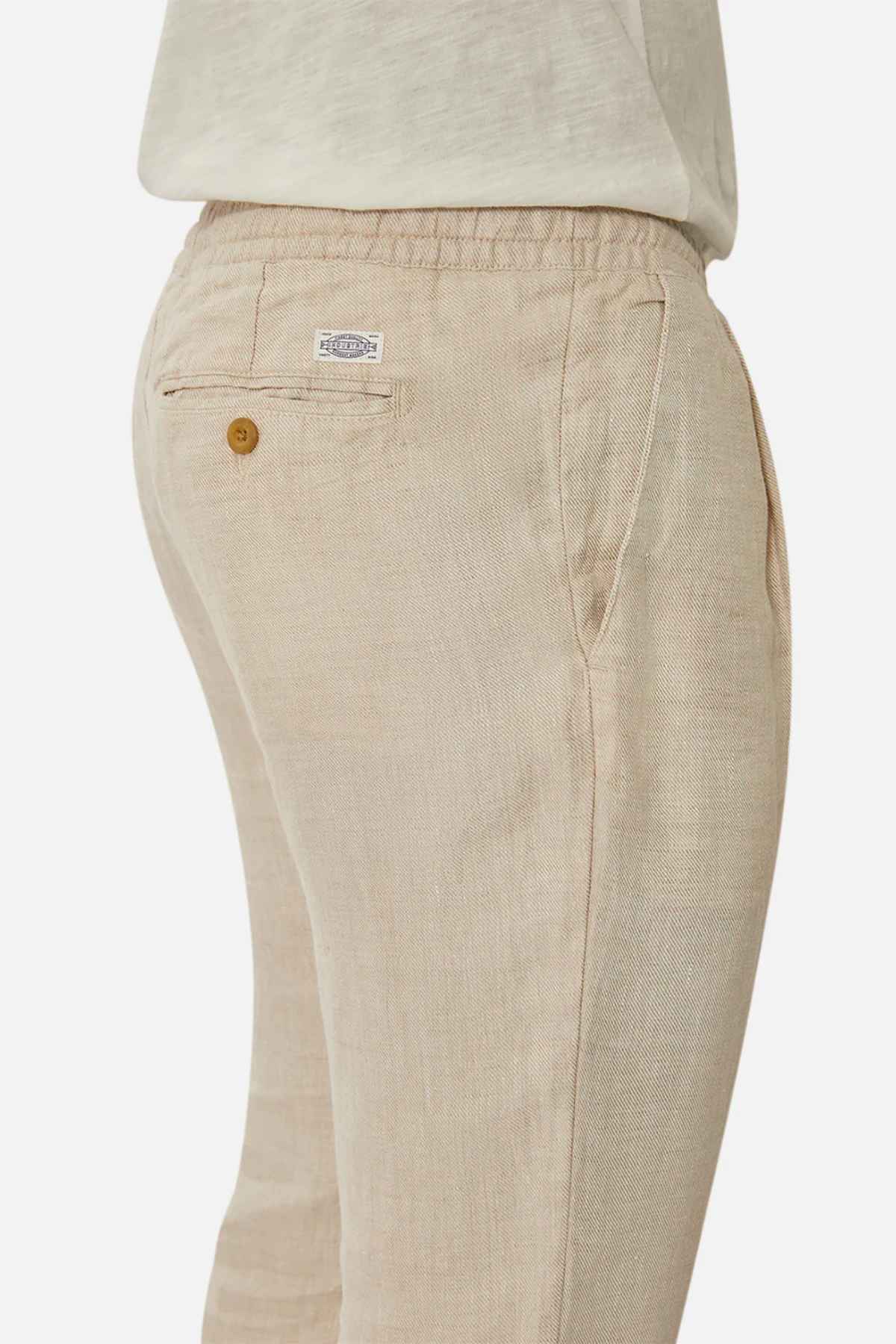 Wearing Industrie The Capri Linen Pant, a wide-leg light beige linen pant featuring an elastic waistband and buttoned back pocket, paired with a light-colored top. This textured fabric offers a casual vibe, ideal for summer.