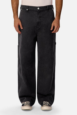 A person wearing Industrie The Carolina Carpenter Pant in black with side pockets, a white shirt tucked in, and white sneakers, standing against a plain light background.
