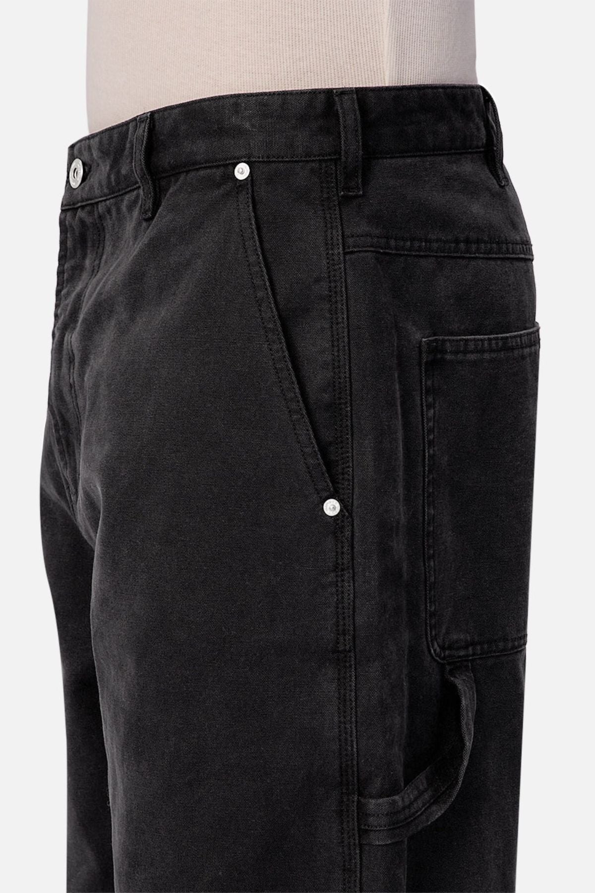 A close-up side view of a person wearing Industrie The Carolina Carpenter Pant, featuring black fabric, silver rivets, a large side pocket, and visible waistband. These relaxed fit pants have a faded look. The top of a beige shirt is also shown.