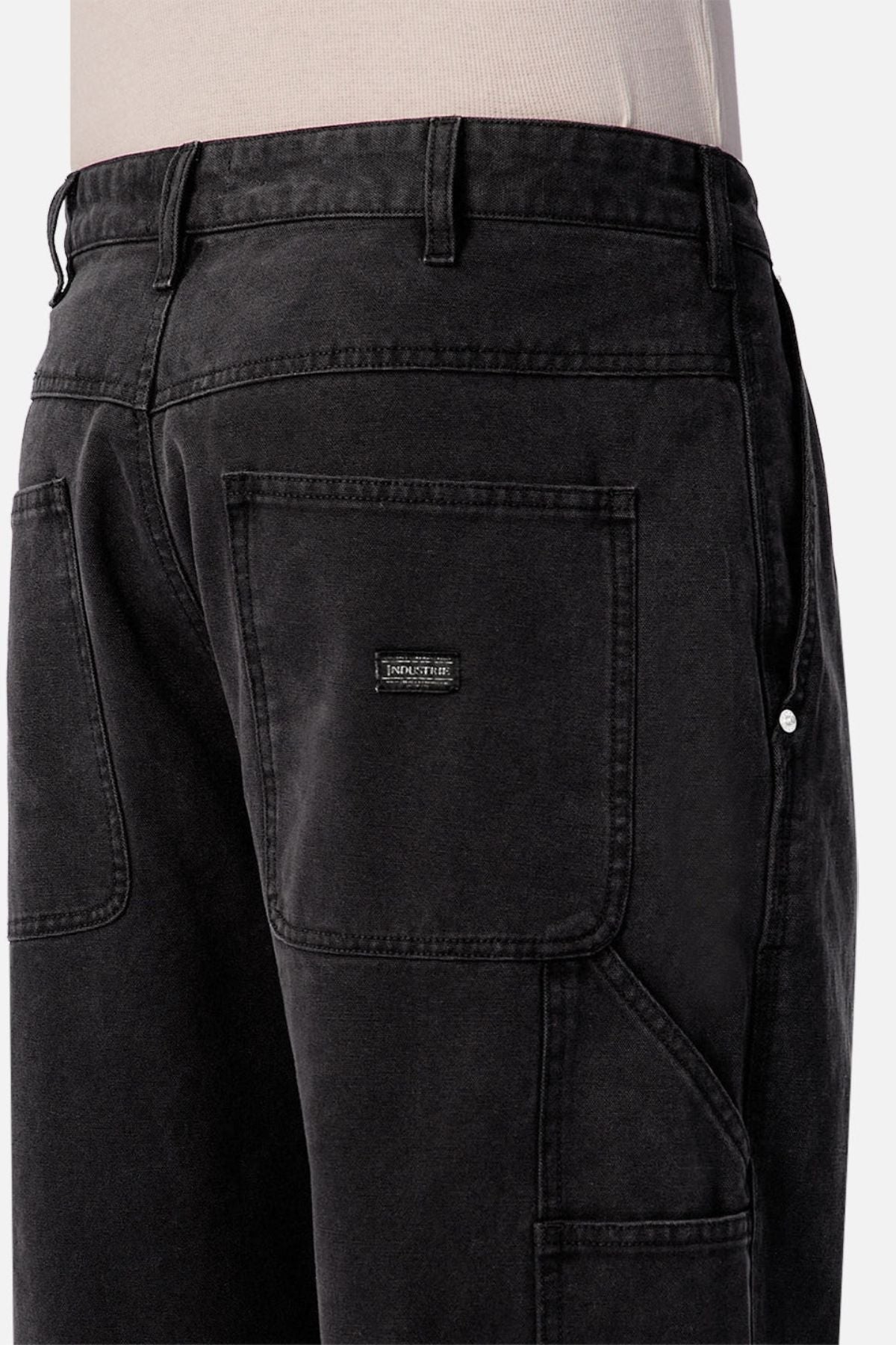 A close-up of someone wearing Industrie The Carolina Carpenter Pant in black denim cotton, showcasing large square pockets, visible belt loops, and a small rectangular black patch on one pocket.