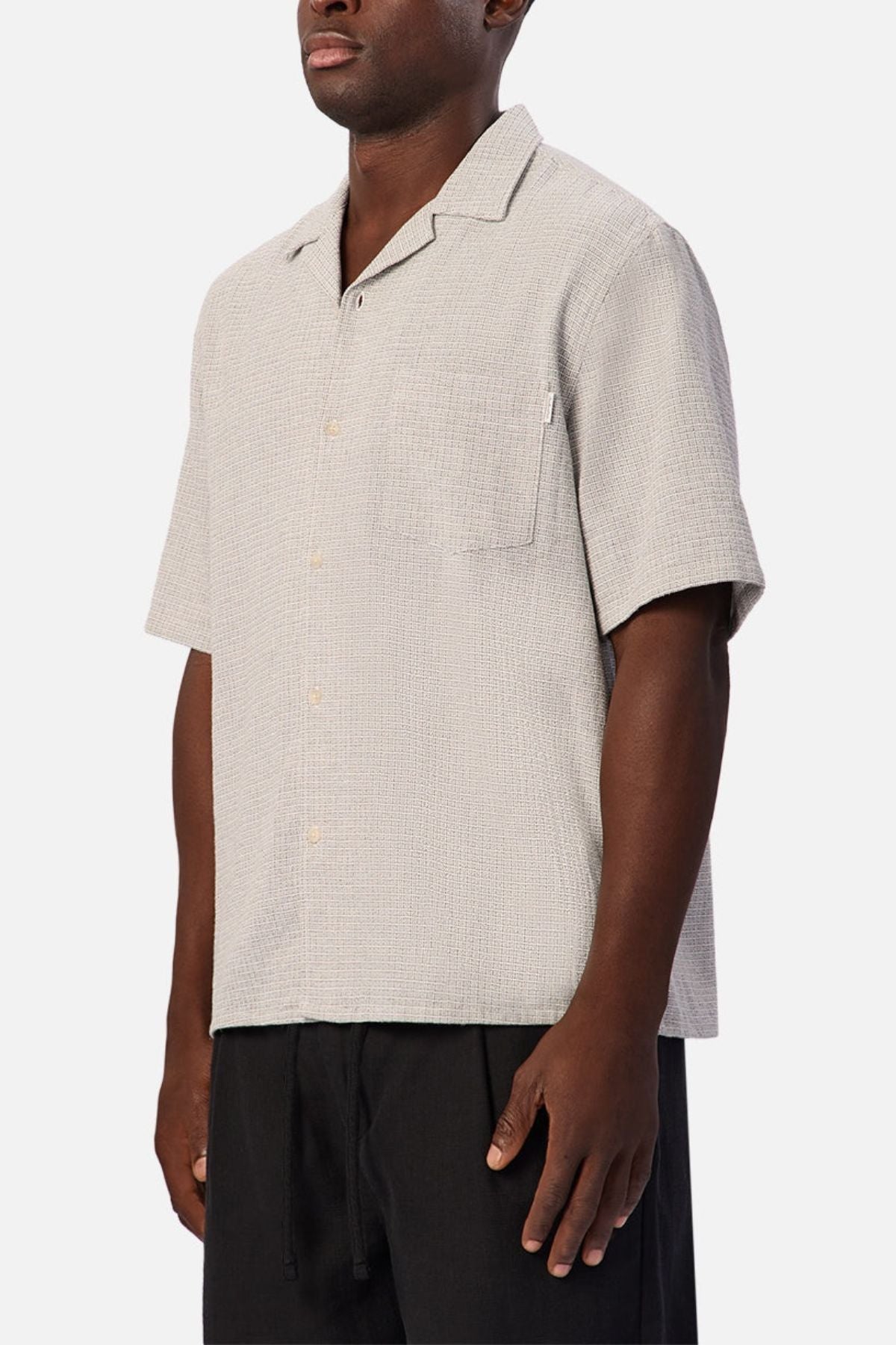 A person wearing the Industrie The Check Marbella Short Sleeve Shirt—a light beige, short sleeve casual shirt with a single chest pocket—paired with black pants, standing against a plain white background.