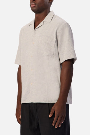 A person wearing the Industrie The Check Marbella Short Sleeve Shirt—a light beige, short sleeve casual shirt with a single chest pocket—paired with black pants, standing against a plain white background.
