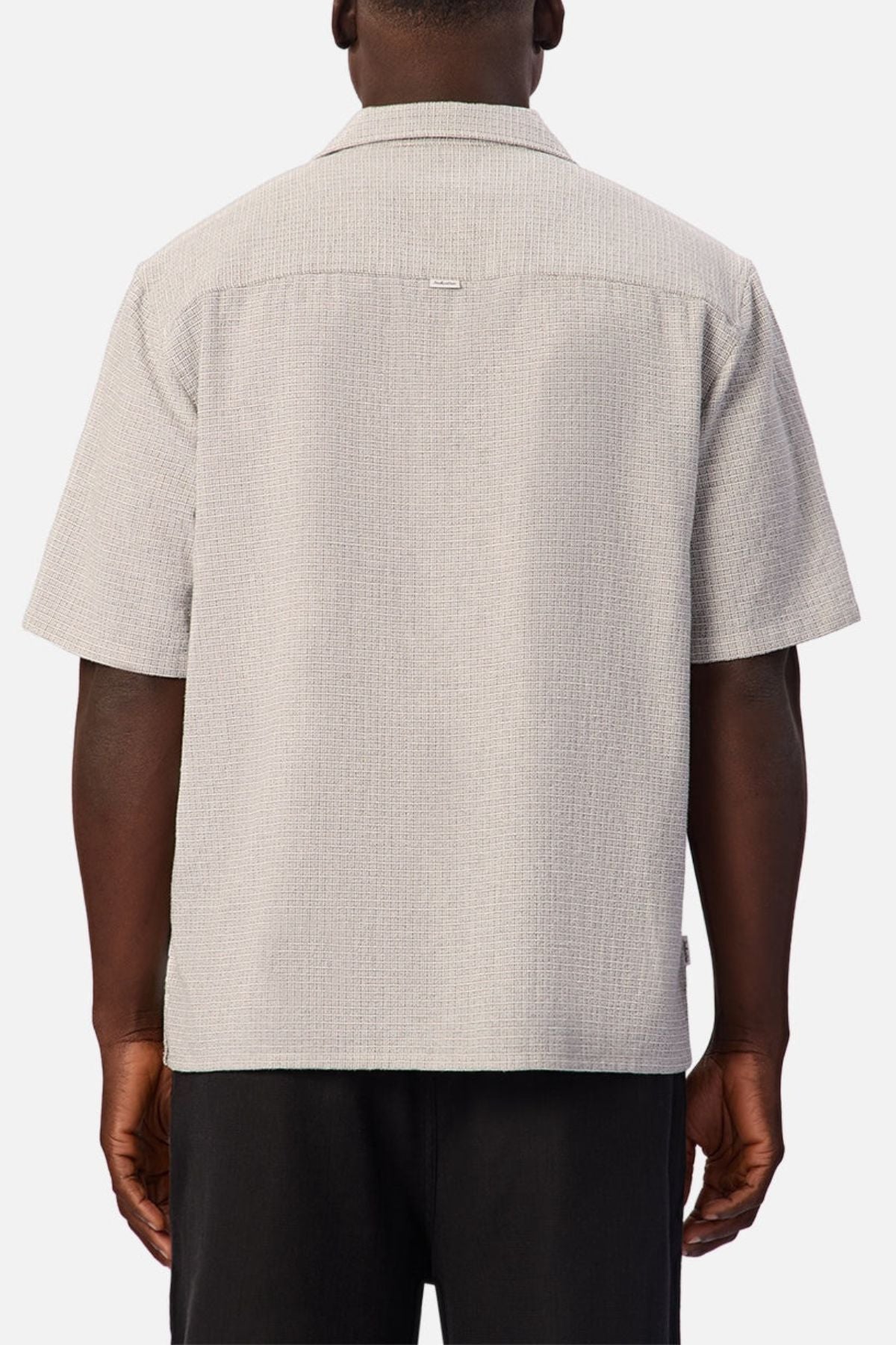 A person stands facing away, wearing the Industrie The Check Marbella Short Sleeve Shirt in light gray textured fabric paired with black pants. The casual shirt features a relaxed fit, a straight hem, and a seam across the upper back. The background is plain.