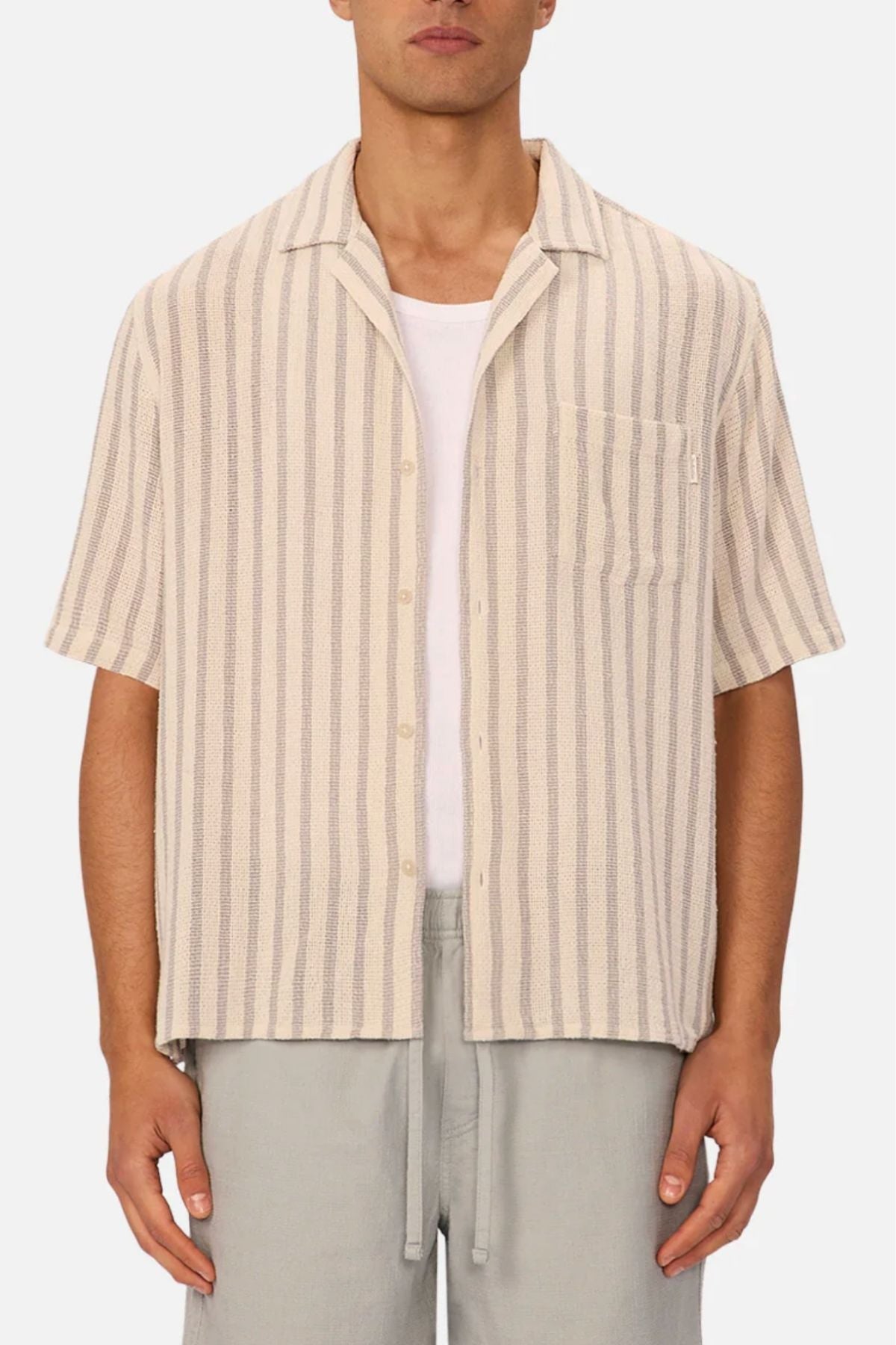 A person wearing the Industrie The Cresta Short Sleeve Shirt in light beige with vertical stripes over a white t-shirt and light gray pants, standing against a plain background.
