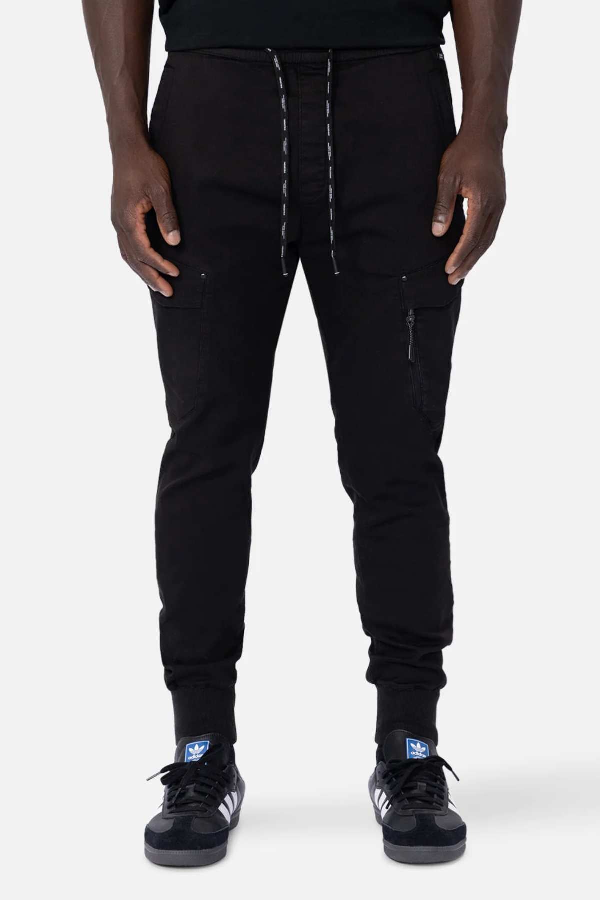 In black Industrie The Defender Combat Pant joggers with drawstrings and thigh zipper pockets, a person stands poised, embodying an active lifestyle. Complementing the look are black sneakers with white stripes and blue logos on the tongues, against a plain white background.