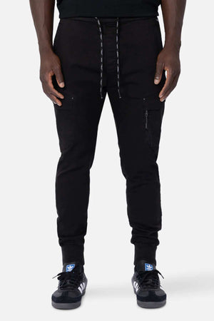 In black Industrie The Defender Combat Pant joggers with drawstrings and thigh zipper pockets, a person stands poised, embodying an active lifestyle. Complementing the look are black sneakers with white stripes and blue logos on the tongues, against a plain white background.