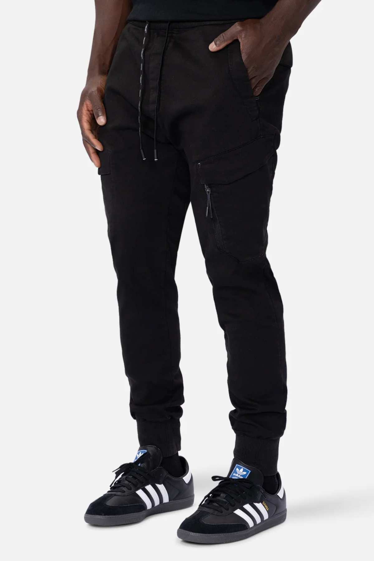 A person is wearing black Industrie The Defender Combat Pants with a drawstring waistband and side pockets, ideal for an active lifestyle. They're also sporting black Adidas sneakers with white stripes, standing confidently against a plain white background.