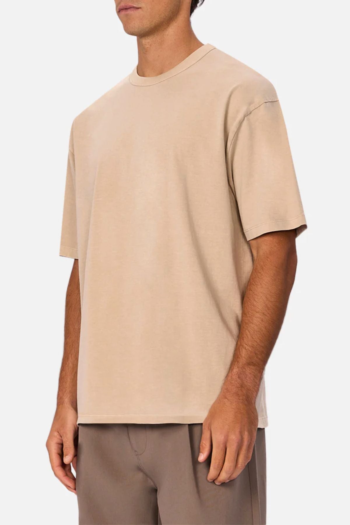 A man wears an oversized beige Industrie The Del Sur Tee made from heavyweight fabric with light brown pants, standing against a plain white background. His face is partially out of frame.