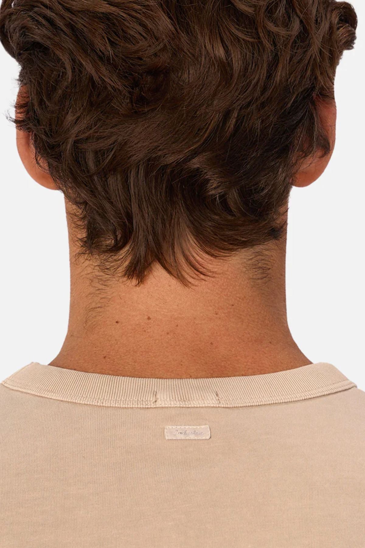 Back view of a person with short, wavy brown hair wearing the Industrie The Del Sur Tee in beige, highlighting its heavyweight fabric and small collar tag with focus on neck and shoulders.
