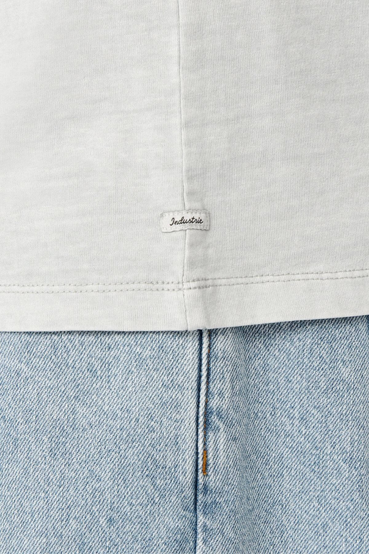 A close-up of a light gray fabric with an "Industrie" label near the seam highlights the oversized 100% cotton Industrie The Del Sur Tee, offering enhanced comfort and style. Below is light blue denim suggesting jeans.
