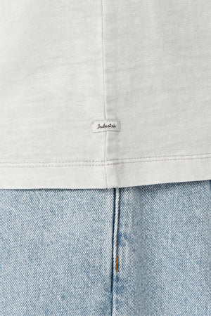 A close-up of a light gray fabric with an "Industrie" label near the seam highlights the oversized 100% cotton Industrie The Del Sur Tee, offering enhanced comfort and style. Below is light blue denim suggesting jeans.