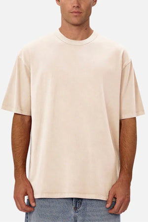 A man in the Industrie The Del Sur Tee, an oversized plain beige t-shirt, paired with light blue jeans and standing against a light gray background.
