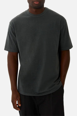A person in an oversized, short-sleeve, dark grey Industrie The Del Sur Tee and black pants stands against a plain light background, with their face partially visible.