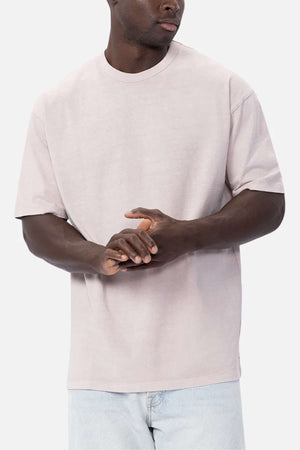 A person is standing against a plain background, wearing the Industrie The Del Sur Tee, an oversized fit T-shirt in light beige made of 100% cotton, paired with light blue jeans. They have their hands clasped together and their face turned slightly to the side.