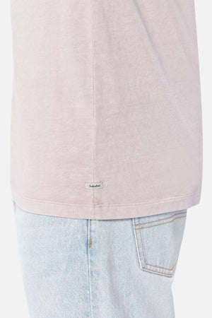 A person wearing a light pink Industrie The Del Sur Tee and light blue jeans. The focus is on the lower part of the 100% cotton t-shirt, which has a small tag with text on the side seam, and the upper part of the jeans, showing a back pocket with visible stitching.